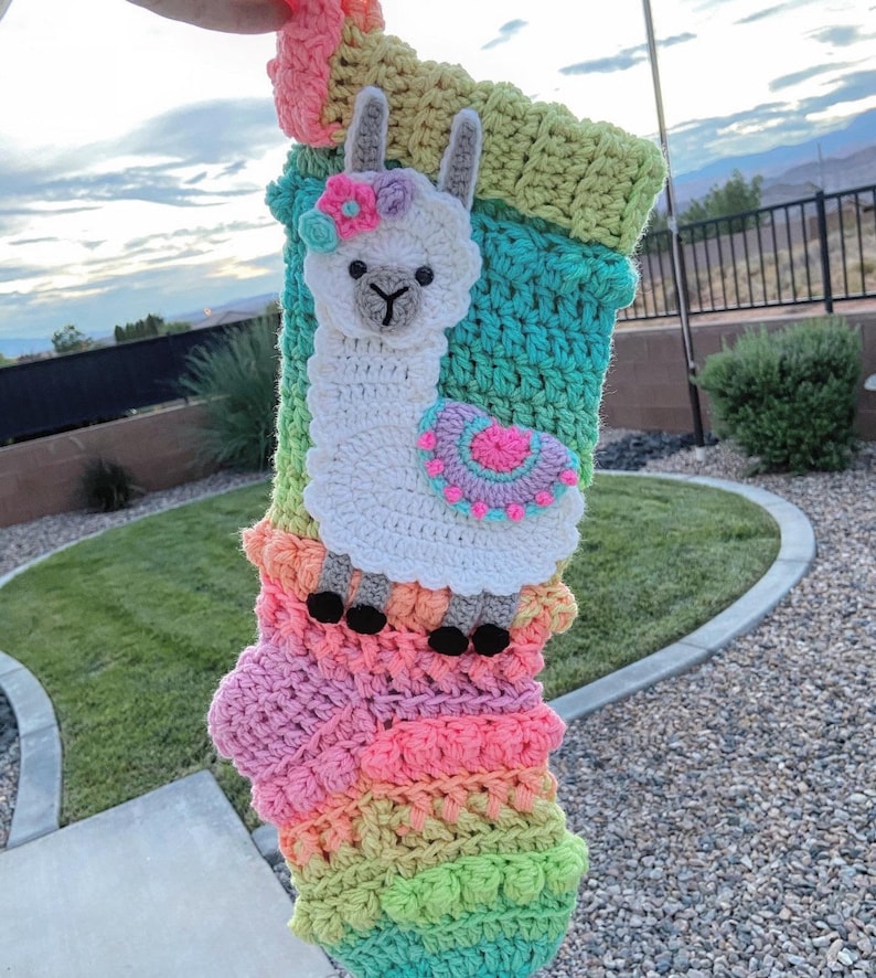 May include: A colorful crochet stocking with a white llama applique. The llama has a pink flower on its head and a pink and purple circle on its side. The stocking is made with a variety of colors, including pink, green, blue, and yellow.