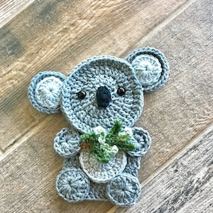 May include: A crocheted koala holding a green leaf. The koala is grey with black eyes and a black nose. The koala is sitting on a wooden surface.