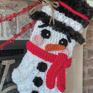 Peut inclure: Chaussette de Noël en crochet blanc en forme de bonhomme de neige avec un chapeau en fourrure noire, une écharpe rouge et des yeux en boutons noirs. La chaussette est accrochée à une cheminée en briques.