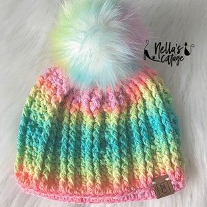 Peut inclure: Bonnet en crochet arc-en-ciel avec un pompon blanc et pastel moelleux. Le bonnet a une étiquette en cuir marron avec les lettres "NC" dessus.