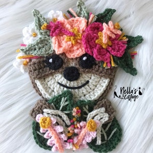 May include: A crocheted sloth wearing a flower crown with pink, peach, and white flowers. The sloth is holding a bouquet of pink, peach, and white flowers. The background is a white fuzzy surface. "Nella's Cottage" is written in the bottom right corner of the image.