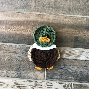 Op de afbeelding: Een gehaakt eenden ornament met een groene kop, bruin lichaam en gele snavel. De eend zit op een houten oppervlak.