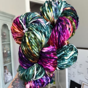 May include: A large skein of hand-dyed yarn in vibrant colors. The yarn is a mix of teal, purple, pink, orange, and yellow. The thick yarn is held together with white string. The yarn is likely for knitting or crocheting projects.