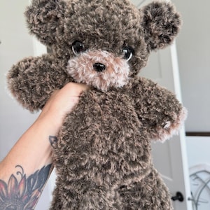 May include: A handmade teddy bear with a fluffy, textured brown body and a lighter brown muzzle. The bear has large, dark eyes and a black nose. The bear is being held up in front of a white background.