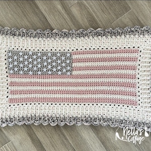 May include: A crocheted American flag with a white background, pink and gray stripes, and a gray star field. The flag is framed with a white and gray scalloped border.