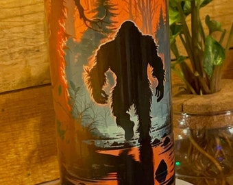 Può includere: Un bicchiere in acciaio inossidabile con un design nero e arancione che presenta una silhouette di Bigfoot che cammina attraverso una foresta con una luna piena sullo sfondo.