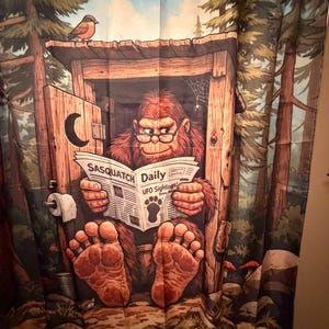 May include: A whimsical shower curtain featuring a cartoon Bigfoot reading a newspaper titled "Sasquatch Daily". The curtain has a forest backdrop with a bird perched on the outhouse roof. The Bigfoot wears glasses and has large feet.