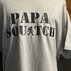 May include: Gray t-shirt with black text that reads "Papa Squatch" with a silhouette of Bigfoot in between the words.