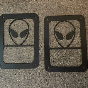 May include: Two black metal alien head cutouts. Each cutout has a square frame with an alien head design in the center. The alien head has large eyes and a rounded head.
