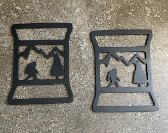 May include: Two black metal wall decor pieces shaped like a can with a mountain scene and a silhouette of a creature inside.