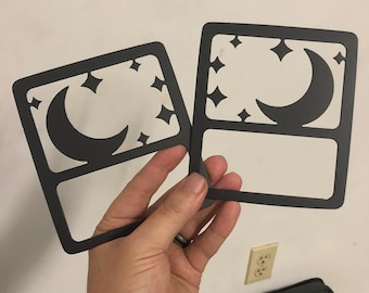 May include: Two black metal cutouts of a crescent moon and stars. The cutouts are in the shape of a square and have a blank space in the center.