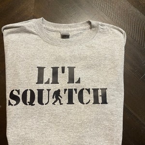 May include: Gray t-shirt with black text that reads "Lil' Squatch" with a small silhouette of Bigfoot walking.