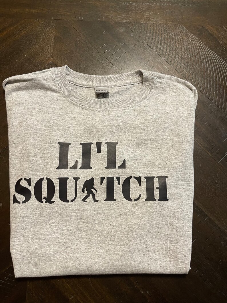 May include: Gray t-shirt with black text that reads "Lil' Squatch" with a small silhouette of Bigfoot walking.
