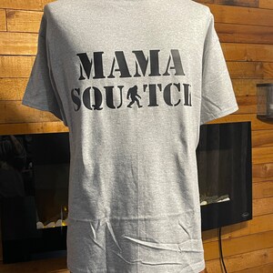 May include: Gray t-shirt with black text that reads "Mama Squatch" with a black silhouette of Bigfoot in the middle of the text.