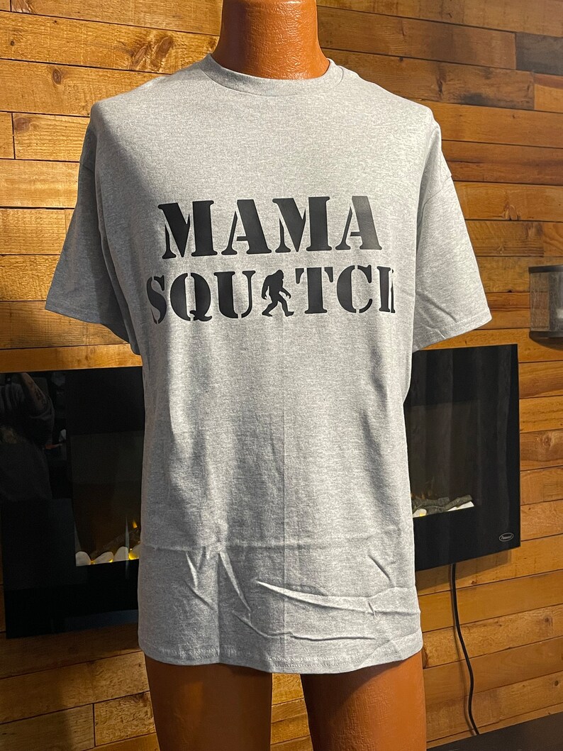 May include: Gray t-shirt with black text that reads "Mama Squatch" with a black silhouette of Bigfoot in the middle of the text.