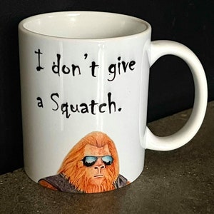 May include: White ceramic coffee mug with a black and white graphic of a sasquatch wearing sunglasses. The text on the mug reads "I don't give a Squatch."