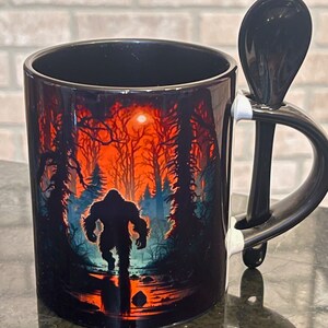 May include: Black ceramic mug with a Bigfoot silhouette walking through a forest scene with a red and orange sunset. The mug includes a black spoon that rests on the handle.