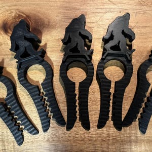 May include: Five black Bigfoot-shaped hair clips are arranged on a wooden surface. Each clip features a silhouette of the legendary creature atop a curved, slotted design with a toothed edge, offering a unique and thematic accessory.