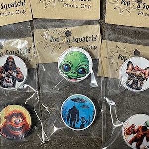 May include: Eight different phone grips with various pop culture characters. The phone grips are round and white with a colorful image on each. The characters include a sasquatch, a creature from the black lagoon, a bigfoot, a french bulldog, and a cartoon monster.