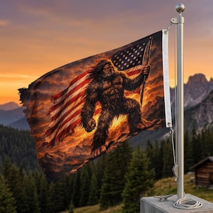 May include: A flag featuring a Bigfoot illustration holding a staff, set against an American flag design with a sunset backdrop. The flag is mounted on a silver pole, with a concrete base, and is waving in the wind.