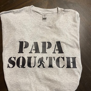 May include: Gray t-shirt with black text that reads "Papa Squatch" with a silhouette of Bigfoot walking.