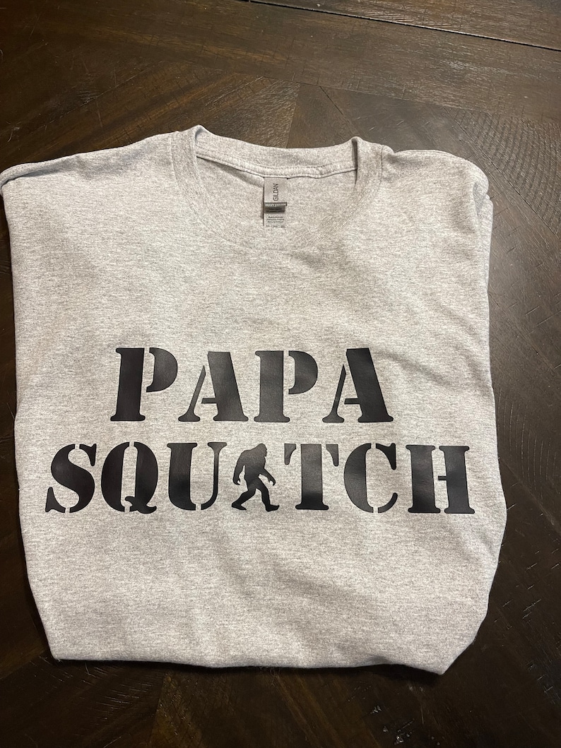 May include: Gray t-shirt with black text that reads "Papa Squatch" with a silhouette of Bigfoot walking.