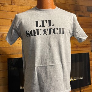 May include: A gray t-shirt with a black graphic that says "Lil' Squatch" with a small image of a Bigfoot walking.