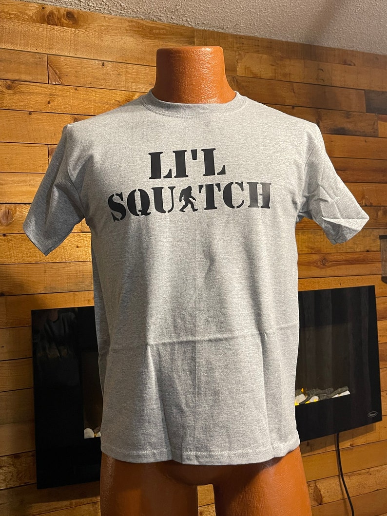 May include: A gray t-shirt with a black graphic that says "Lil' Squatch" with a small image of a Bigfoot walking.