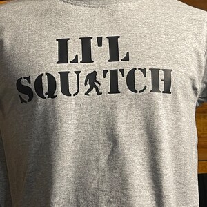 May include: Gray t-shirt with black text that reads "Lil' Squatch" with a small black silhouette of a Bigfoot walking in front of the word "Squatch".