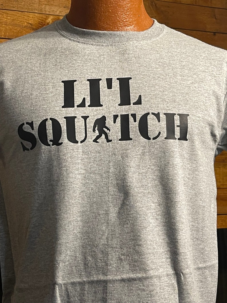 May include: Gray t-shirt with black text that reads "Lil' Squatch" with a small black silhouette of a Bigfoot walking in front of the word "Squatch".