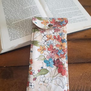 May include: A beige fabric pencil case with a floral print featuring pink, orange, yellow, and blue flowers. The case has a flap with a button closure.
