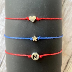 May include: Three red string bracelets with silver charms. The top bracelet has a heart charm, the middle bracelet has a star charm, and the bottom bracelet has a round charm with the letter "M" on it.