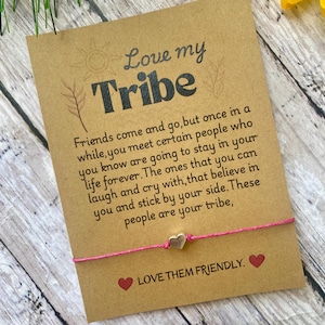May include: A pink string bracelet with two silver heart charms and a card that reads "Love my Tribe" with a quote about friendship. The card has a brown background and a sunflower graphic.