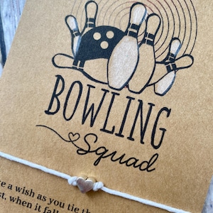 May include: A brown card with a bowling pin graphic and the words "BOWLING Squad". A white string bracelet with a silver heart charm is attached. The card has text that reads "Make a wish as you tie the bracelet on your wrist, when it falls off, the wish you made will soon come true!"