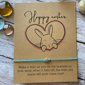 May include: A brown card with the text "Hoppy Easter" and a cartoon bunny rabbit peeking out of a pink heart. The card is tied with a light blue string with a silver star charm. The text "Make a wish as you tie the bracelet on your wrist, when it falls off, the wish you made will soon come true!" is printed on the card.