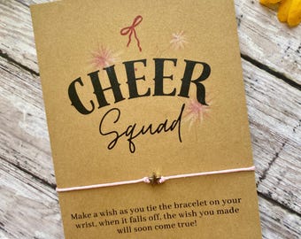 May include: A brown card with the text "CHEER Squad" in black lettering. A pink string bracelet with a gold star charm is attached to the card. The card reads "Make a wish as you tie the bracelet on your wrist, when it falls off, the wish you made will soon come true!"