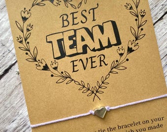 May include: A brown card with a black heart-shaped floral design and the text "BEST TEAM EVER". A white string bracelet with a silver heart charm is attached to the card. The card reads "Make a wish as you tie the bracelet on your wrist, when it falls off, the wish you made will soon come true!"