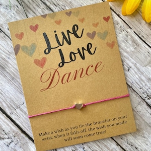 May include: A pink string bracelet with a silver heart charm. The bracelet is tied around a card with the words "Live Love Dance" and a message about making a wish when the bracelet falls off.