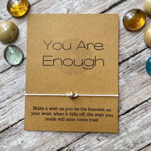 May include: A white string bracelet with a silver star charm. The bracelet comes with a card that reads "You Are Enough" and instructions to make a wish when you tie the bracelet on your wrist. When the bracelet falls off, the wish will come true.