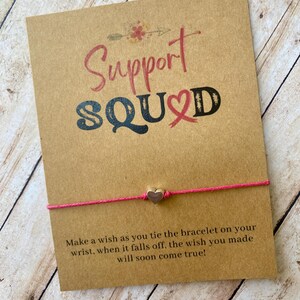 May include: A pink string bracelet with a small silver heart charm is displayed on a brown card. The card has the words "Support SQUAD" in pink and black lettering, with an arrow and floral design. Text at the bottom reads, "Make a wish..."