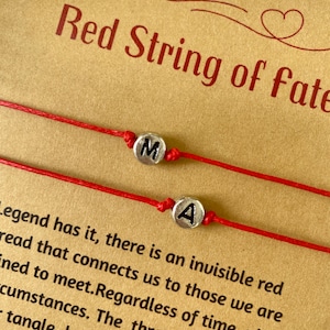 May include: Two red string bracelets with silver charms that say "M" and "A". The bracelets are on a brown background with the text "Red String of Fate" and a legend about the red string of fate.