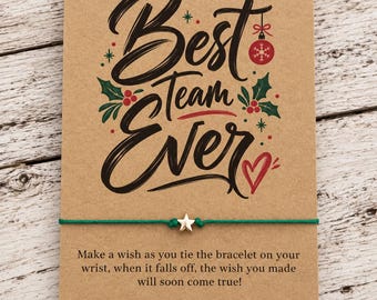 May include: A green string bracelet with a small gold star charm attached to a rectangular kraft paper card. The card features the text "Best Team Ever" in black script, a red ornament, holly leaves, and a red heart, plus instructional text.