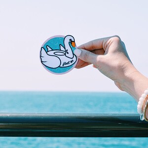May include: A white swan-shaped inflatable pool float sticker with the text "float on" in pink lettering on a blue and white background.