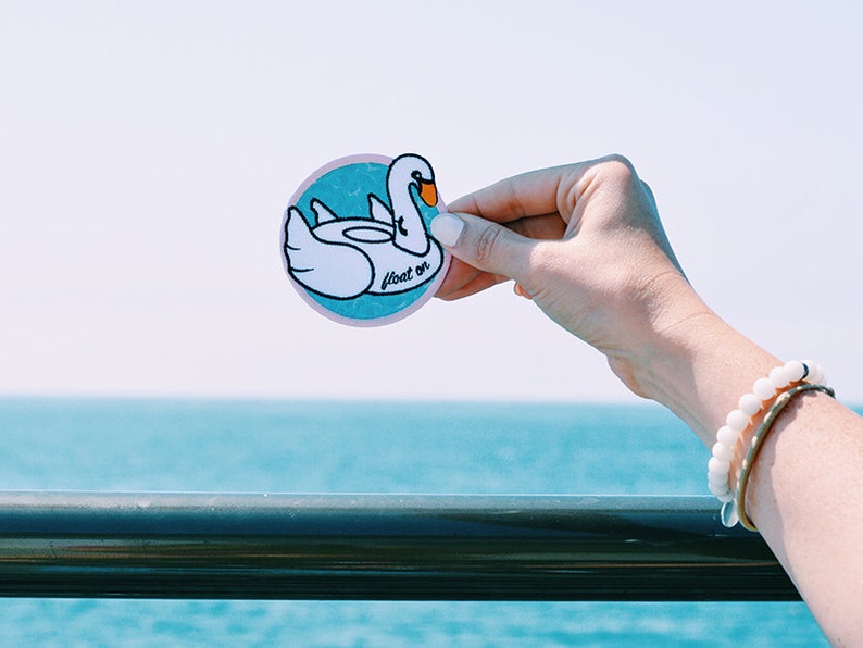 May include: A white swan-shaped inflatable pool float sticker with the text "float on" in pink lettering on a blue and white background.