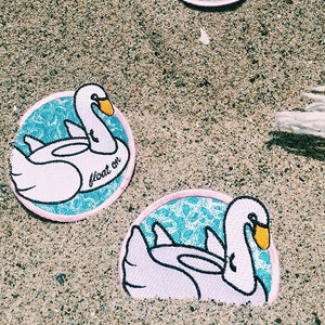 May include: Two embroidered iron-on patches featuring a white swan on a blue and white patterned background. The swan is in a floatie shape and the text "float on" is written on the patch.