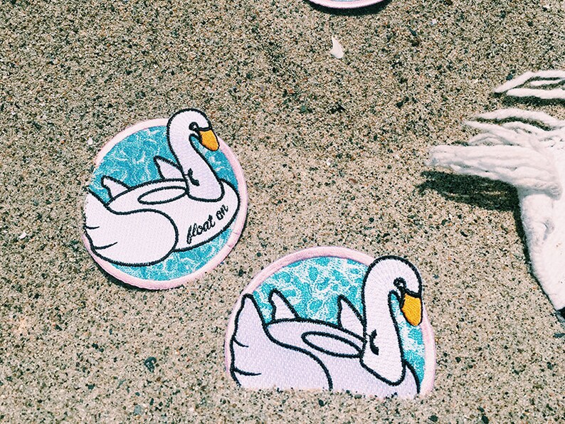 May include: Two embroidered iron-on patches featuring a white swan on a blue and white patterned background. The swan is in a floatie shape and the text "float on" is written on the patch.