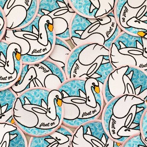 May include: A collection of embroidered iron-on patches featuring white swans on a blue and white water pattern. Each patch has a pink border and the text "float on" in black.