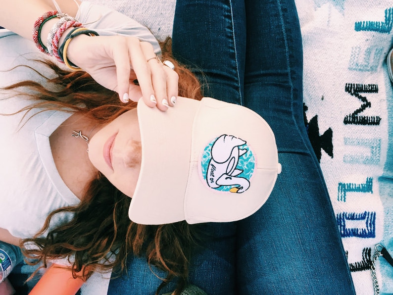 May include: A pink baseball cap with a white swan graphic and the text "float on" in blue. The swan is holding a yellow object in its beak.