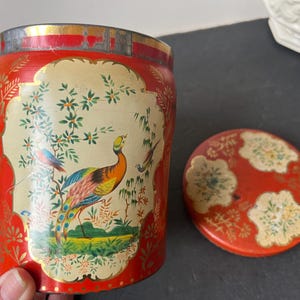 May include: Vintage red and gold tin container with a lid. The container features a painted design of a peacock and other birds amidst floral elements. The lid is red with floral patterns. The container is cylindrical.