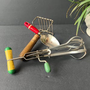 May include: A collection of vintage kitchen utensils, including a hand-crank egg beater with a green and yellow handle, a potato masher, and a spoon. The handles are wood and painted red, green, and yellow.